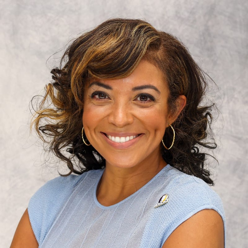 Atasha M. James, smiling warmly in a light blue knit top and gold hoop earrings, against a soft gray studio backdrop